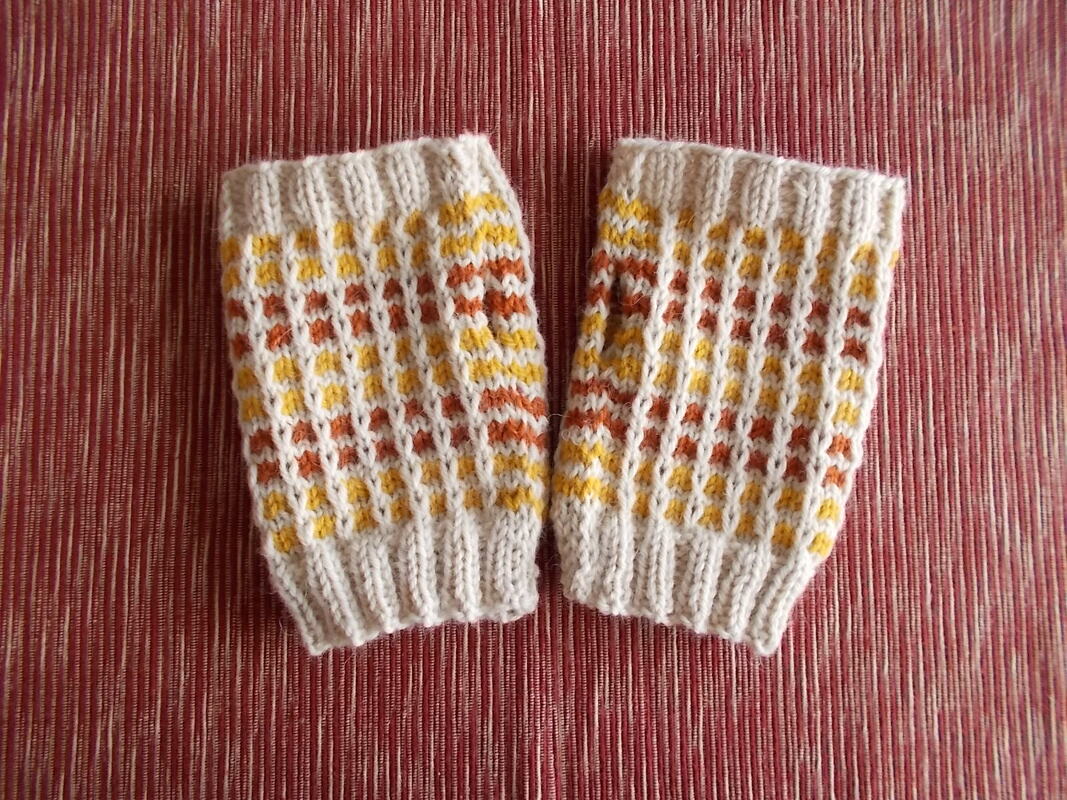 Zwei gestrickte Armstulpen in Creme mit gelben und orangefarbenen Mustern auf rotem, strukturiertem Untergrund.