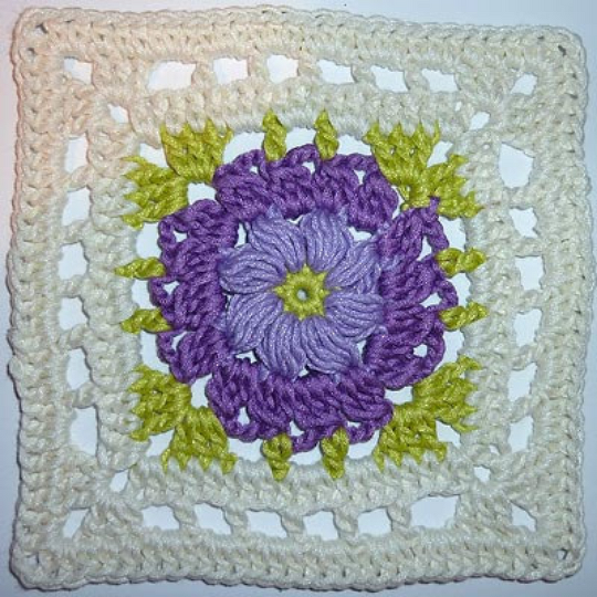 gehäkeltes Granny Square mit Reliefblüte in Lila, grünen Blattakzenten und cremefarbenem Rand