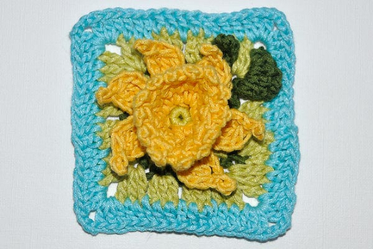 Gehäkeltes Granny Square mit gelber 3D-Blüte, grünen Blättern und türkisfarbenem Rand