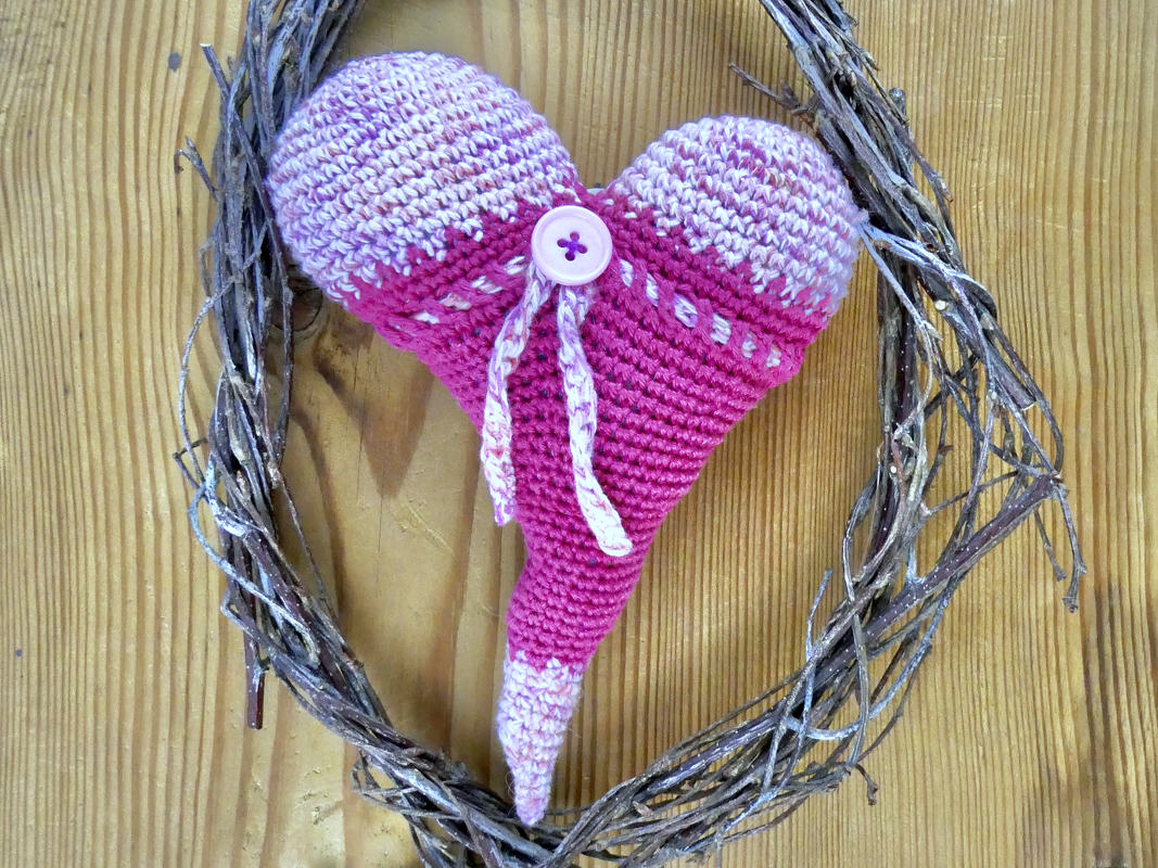 Pink-lila gehäkeltes Herz mit Knopf und Band in Astkranz auf Holz
