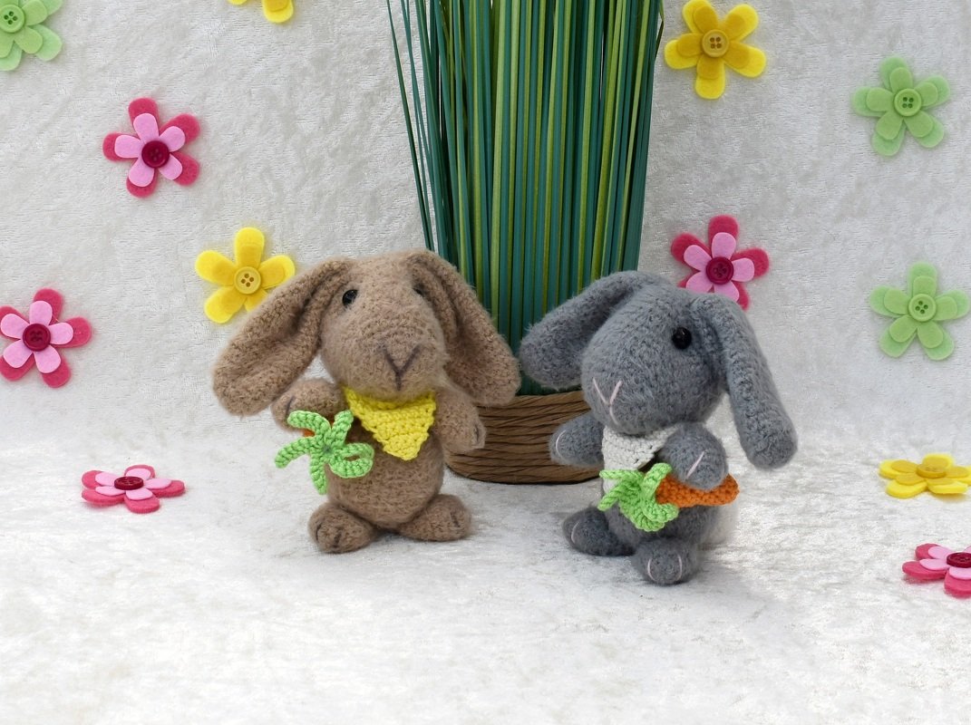 Zwei gehäkelte Hasen (braun und grau) mit Halstüchern, Karotte und Blatt, umgeben von Filzblumen auf hellem Untergrund.