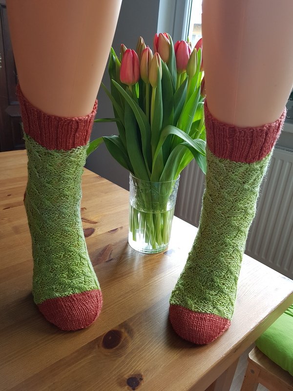 Grüne handgestrickte Socken mit Strukturmuster und terrakottafarbenen Bündchen und Spitze an Schaufensterpuppenbeinen, Tulpen im Hintergrund.