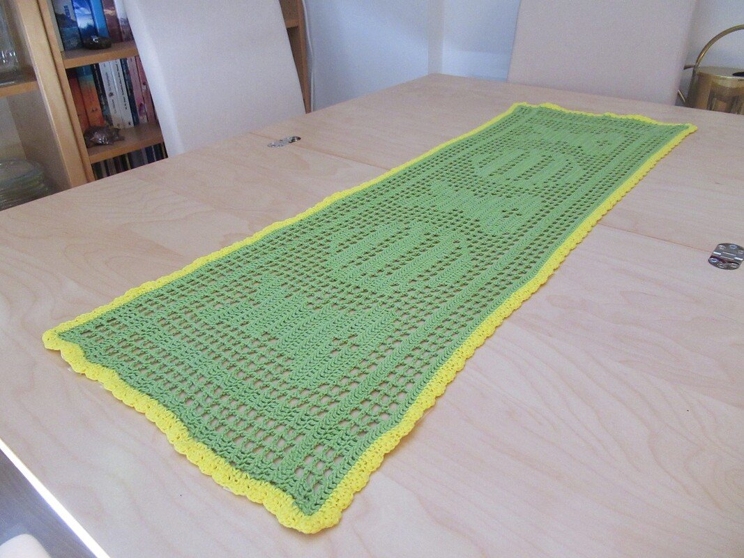 Hellgrüner gehäkelter Tischläufer mit gelbem Wellenrand und eingearbeitetem Hasenmuster auf einem Holztisch.