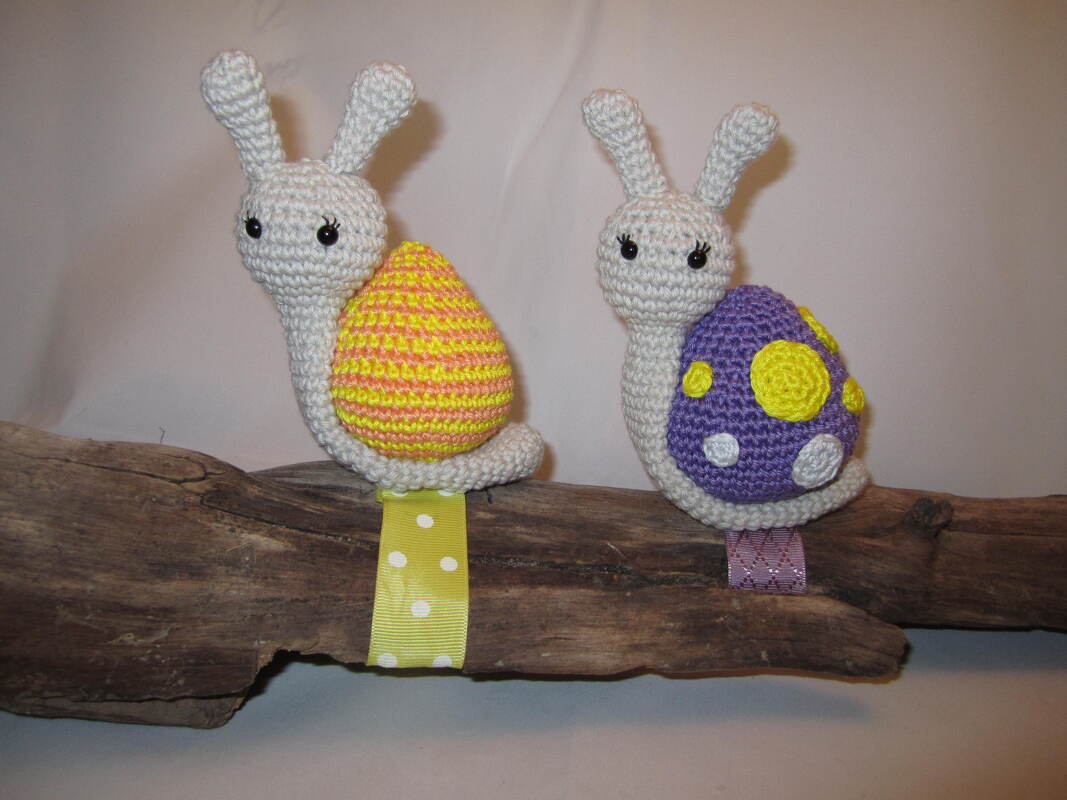 Zwei gehäkelte Amigurumi-Schnecken mit cremefarbenen Körpern, eine mit gelb-orangem gestreiftem Panzer, die andere mit violettem Panzer und Punkten, auf Treibholz