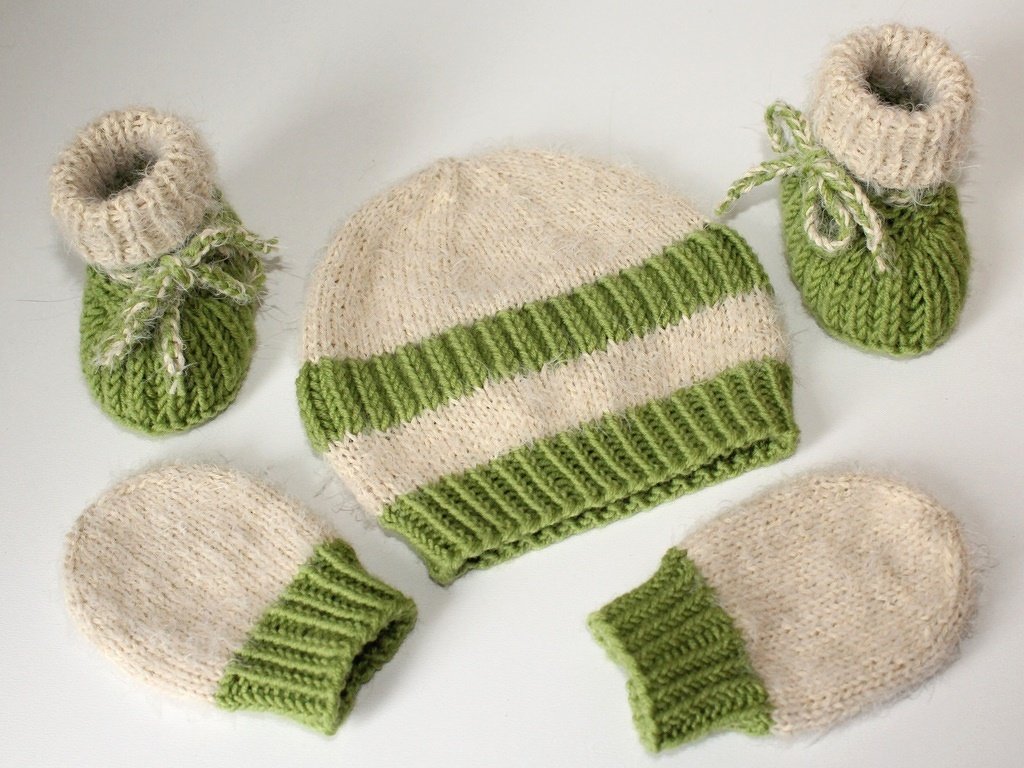 Gestrickte Baby-Mütze, zwei Fäustlinge und ein Paar Babyschuhe in Creme und Grün auf hellem Hintergrund
