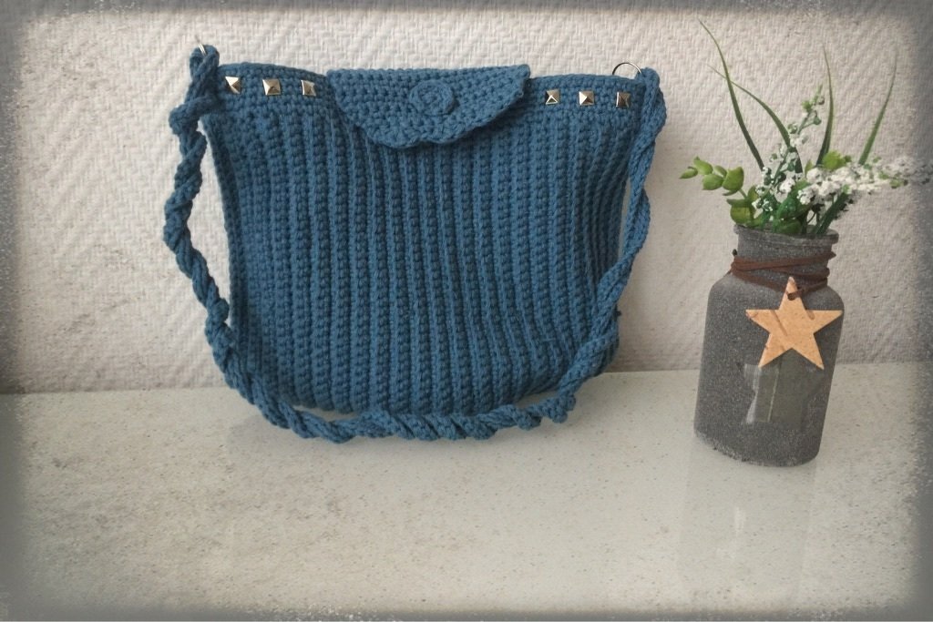 Blaue gehäkelte Umhängetasche mit Nieten an der Klappe und gedrehtem Träger, daneben ein dekoriertes Glas mit Blumen