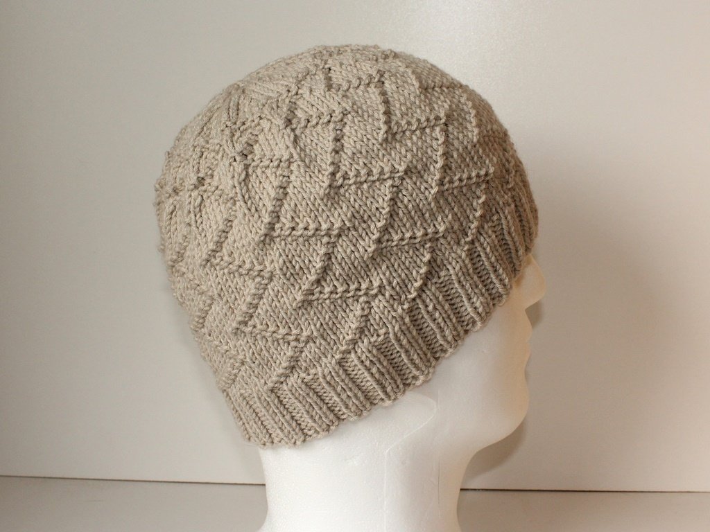 Beige Strickmütze mit Rautenmuster auf weißem Schaufensterkopf, seitliche Ansicht.