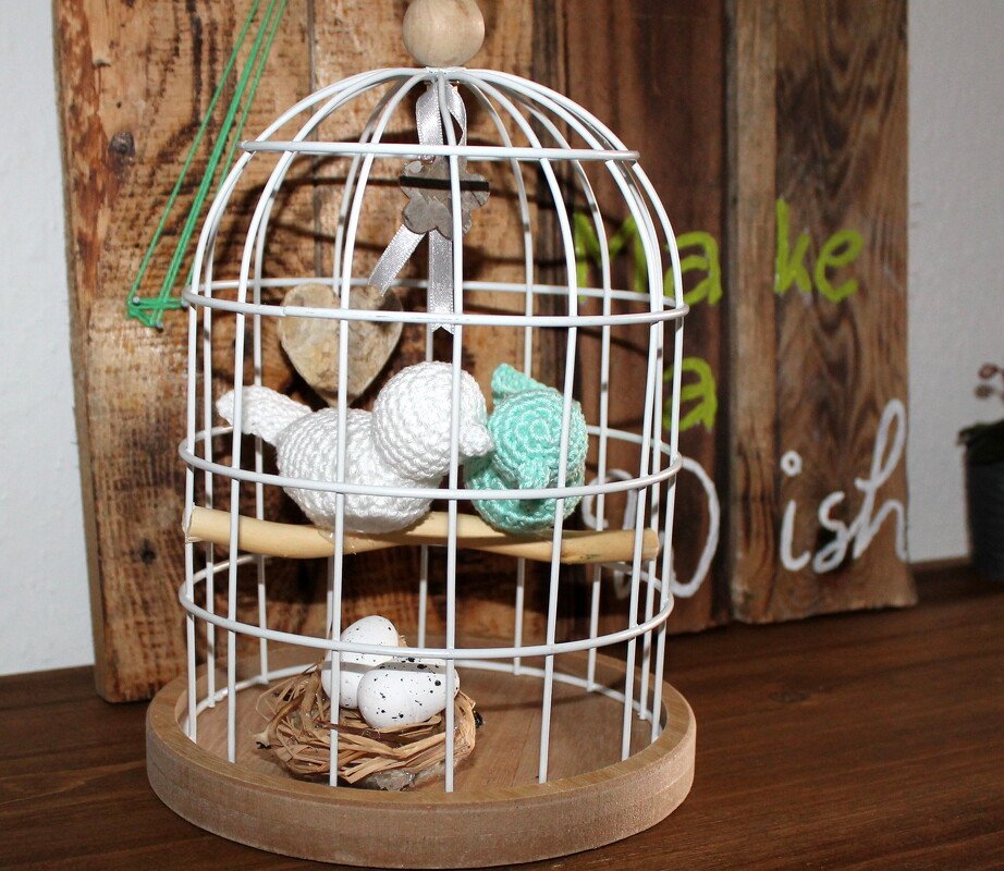Weißer Draht-Vogelkäfig mit zwei gehäkelten Amigurumi-Vögeln auf einer Holzstange, Nest mit gesprenkelten Eiern und Holzboden.