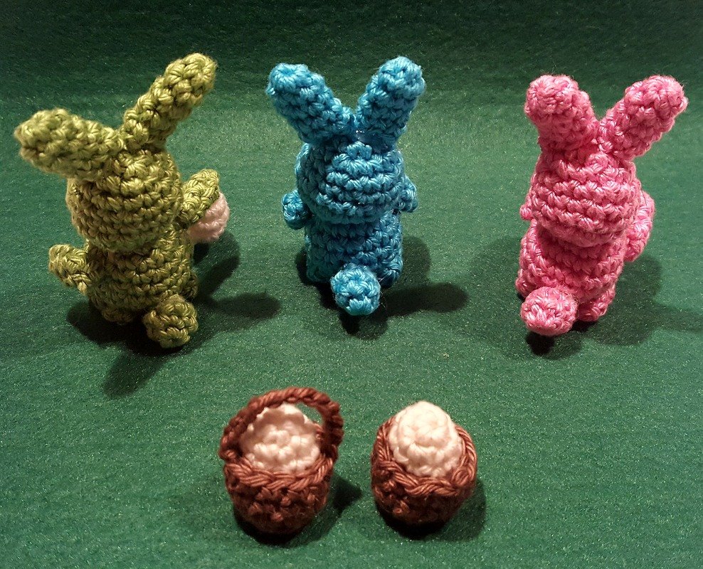 Drei gehäkelte Amigurumi-Hasen (grün, blau, rosa) mit zwei kleinen braunen Körbchen auf grünem Untergrund.