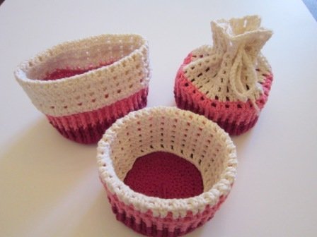 Drei gehäkelte Behälter: ein ovaler Korb, ein runder Korb und ein runder Zugbeutel, alle in Rosa- und Cremefarben.