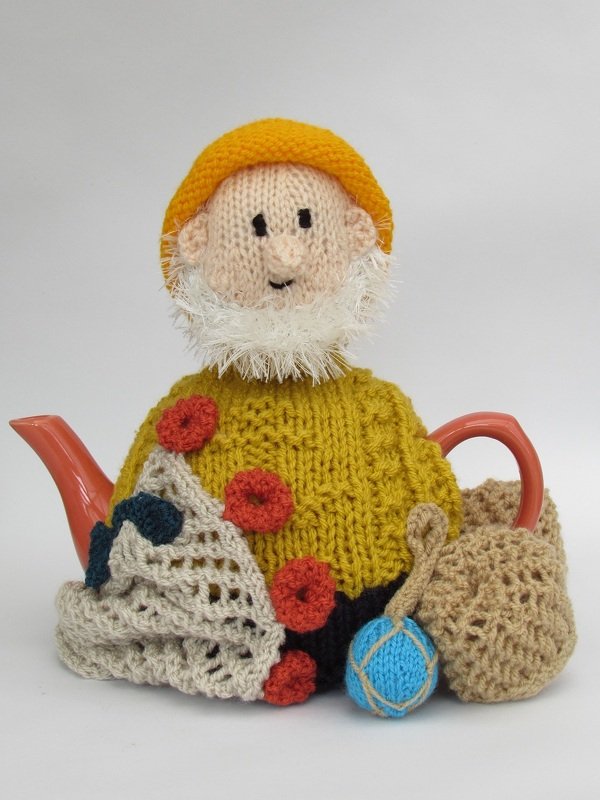 Knitted tea cozy shaped like a fisherman with yellow hat, white beard, mustard-yellow sweater, net overlay, red floats and blue buoy on a teapot.