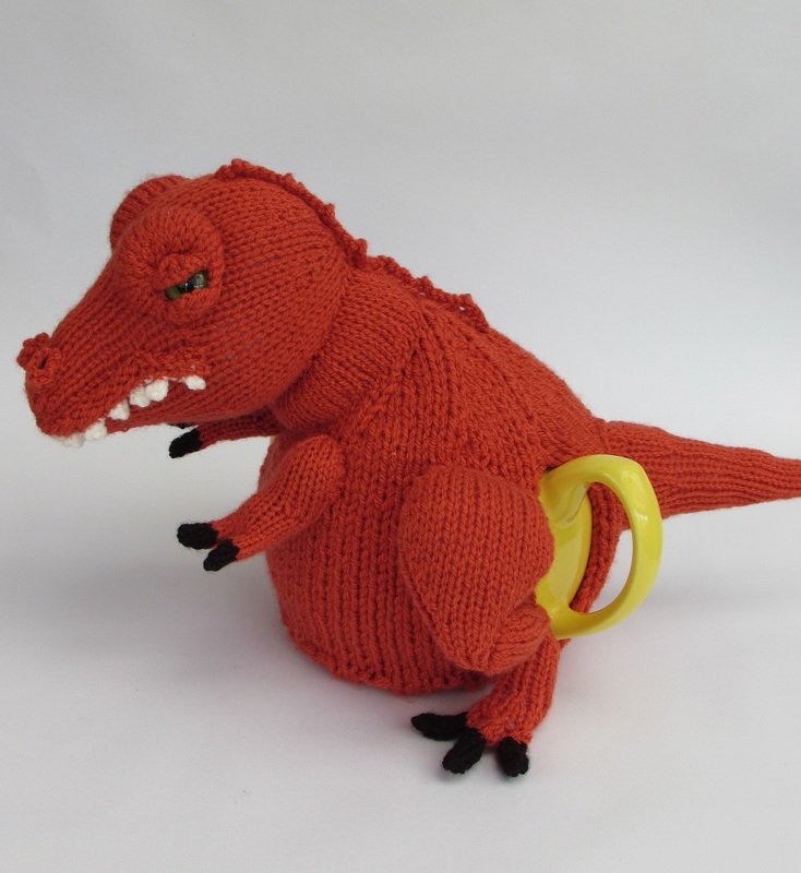 Red hand-knitted T-Rex tea cozy with green eye, white teeth and black claws covering a teapot with a yellow handle