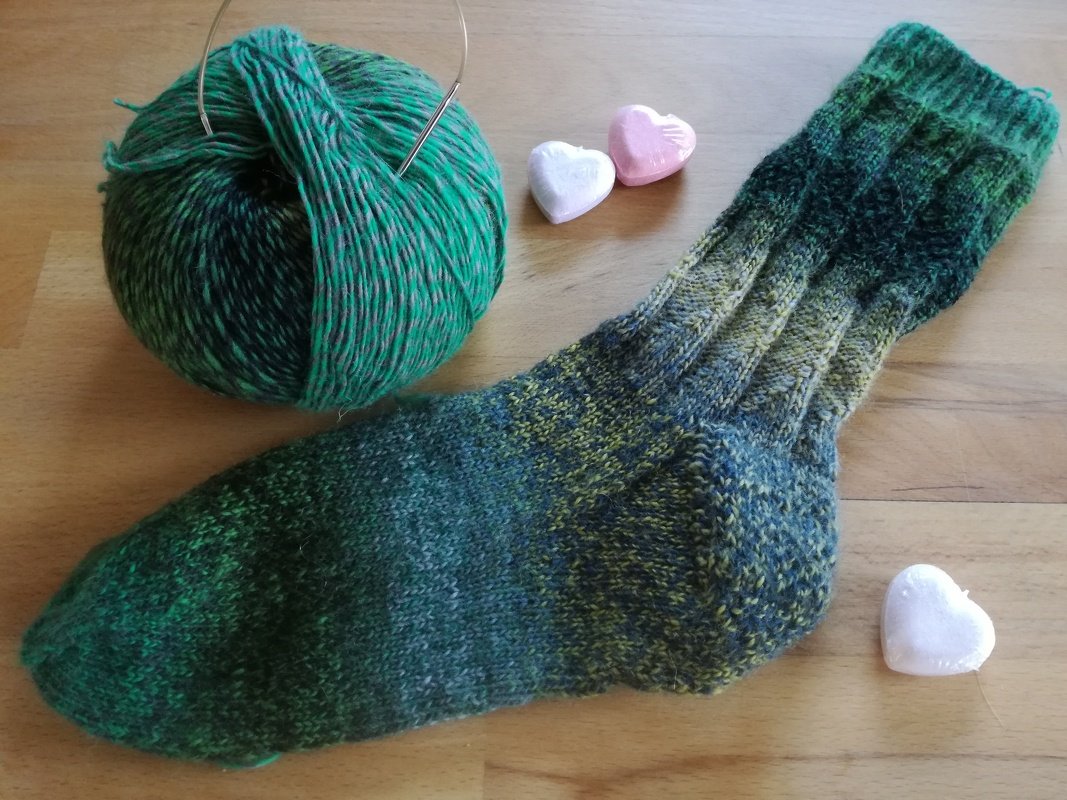 Handgestrickte grün-gelbe Socke neben Garnknäuel, Rundstricknadel und kleinen Herz-Dekorationen auf Holz.