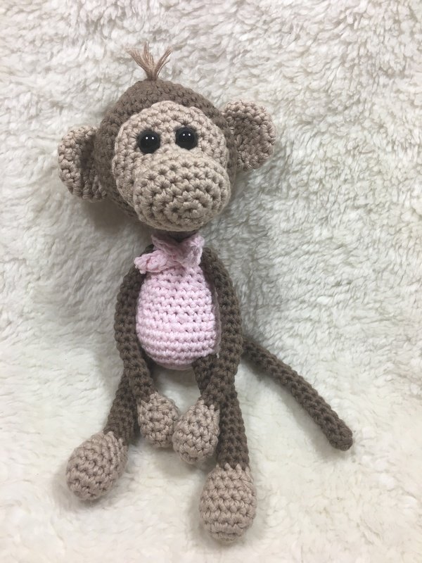 Handgehäkelter brauner Amigurumi-Affe mit rosafarbenem Bauch und Halstuch auf hellem Plüsch