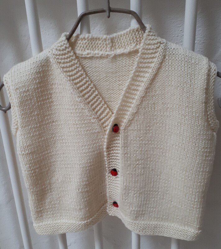 Cremefarbene gestrickte Kinderweste mit V-Ausschnitt und drei roten Knöpfen auf einem Bügel