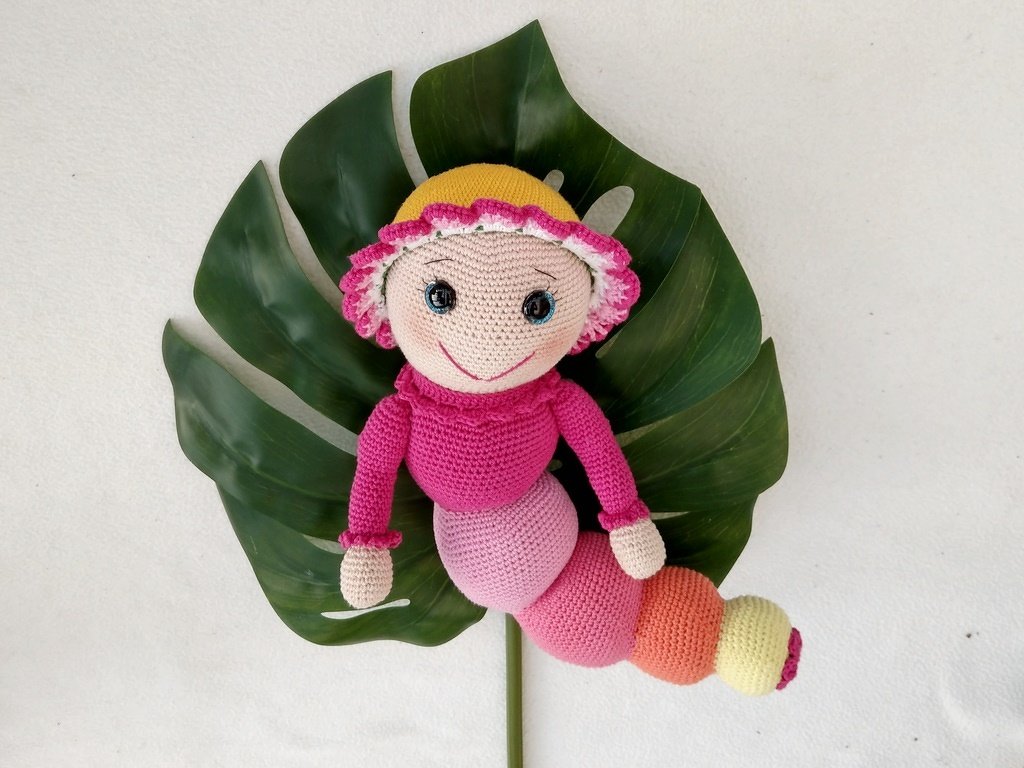 Gehäkelte Raupenpuppe in Pink und Rosa mit gelben Akzenten, liegend auf einem großen grünen Monstera-Blatt