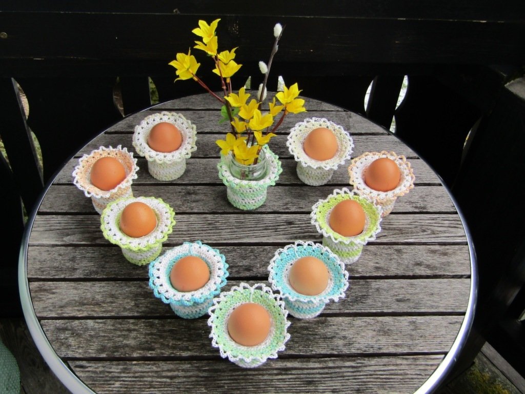 Neun gehäkelte Eierwärmer mit braunen Eiern in Herzform auf einem Holztisch, Vase mit gelben Frühlingsblüten