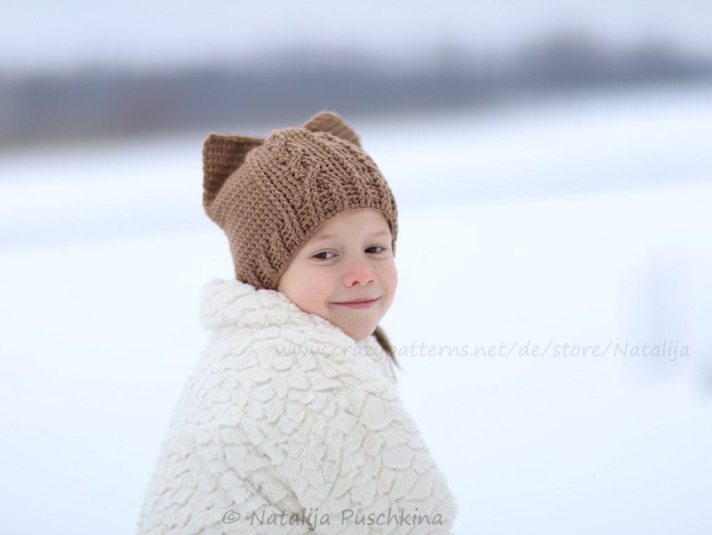 Kind mit brauner gehäkelter Mütze mit Ohren und weißer Jacke im Schnee