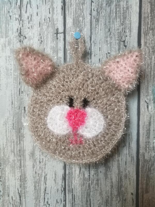 Gehäkelter Katzenkopf in hellbraun mit rosa Ohren und Nase, an einer hellen Holzwand befestigt.