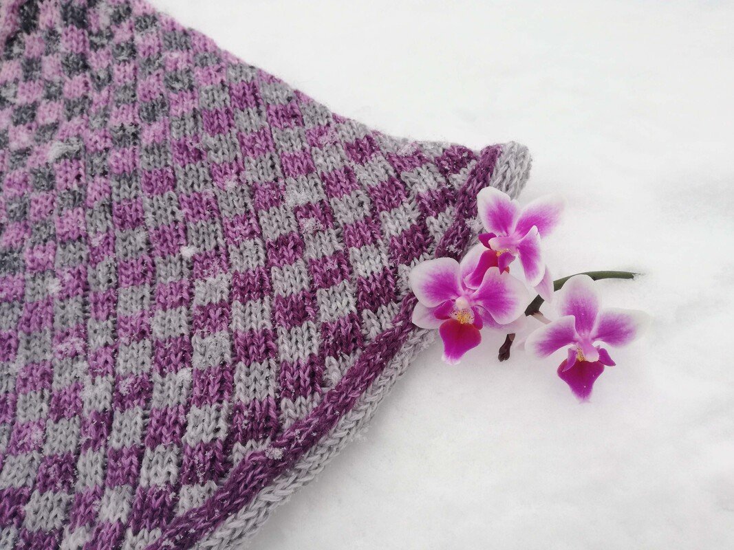 Gestrickter Loop in Schachbrettmuster in Grau und Violett mit drei Orchideenblüten daneben
