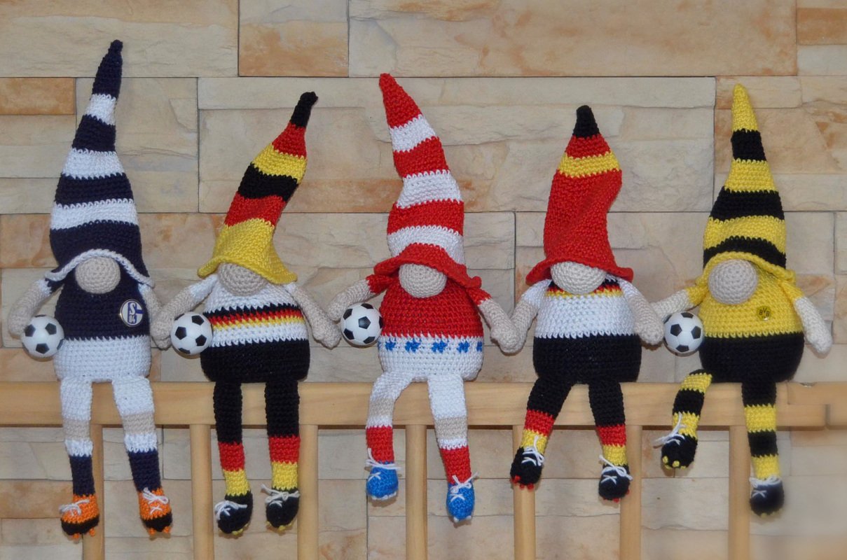 Fünf gehäkelte Zwerge in Fußballfarben auf einer Holzleiste, jeder mit einem kleinen Fußball.