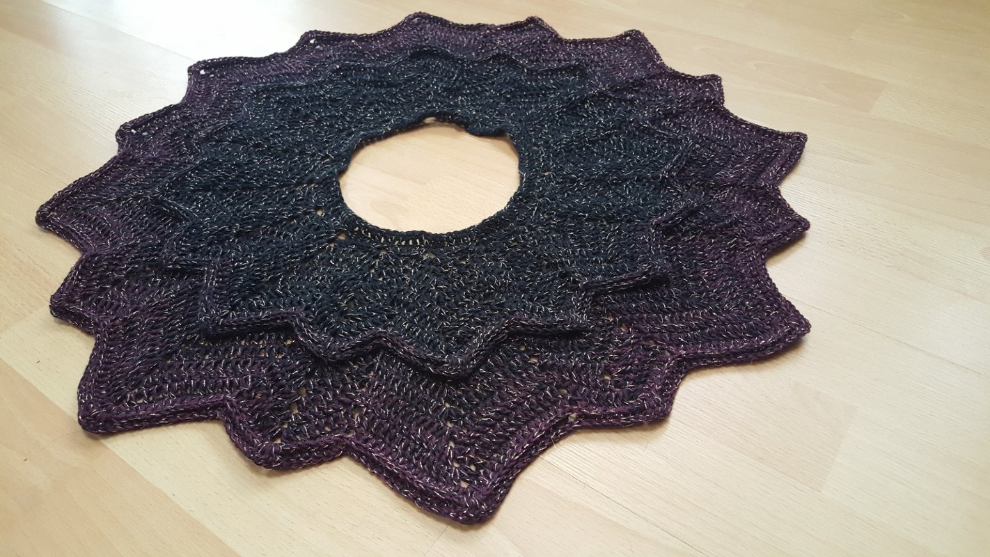 Runder gehäkelter Kragenschal in Schwarz und Violett mit Glitzergarn, flach auf hellem Holzboden.