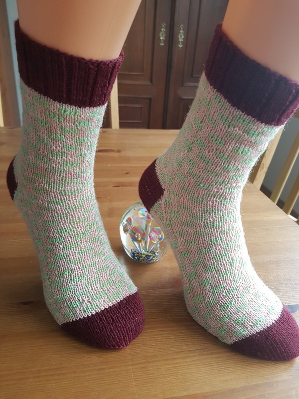 Hand-knitted socks with pale pink and mint-green flecked body and burgundy cuffs, heels, and toes on a wooden table.