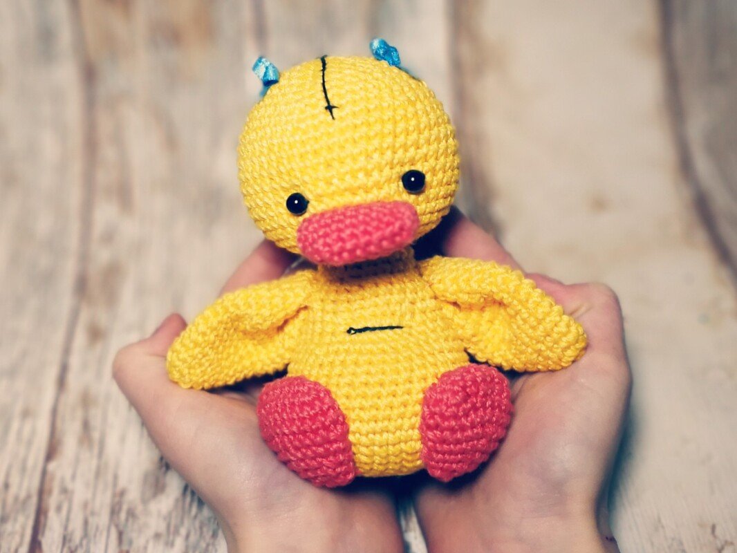 Gelbe Amigurumi-Ente mit rosa Schnabel und Füßchen in Händen vor hellem Holzhintergrund