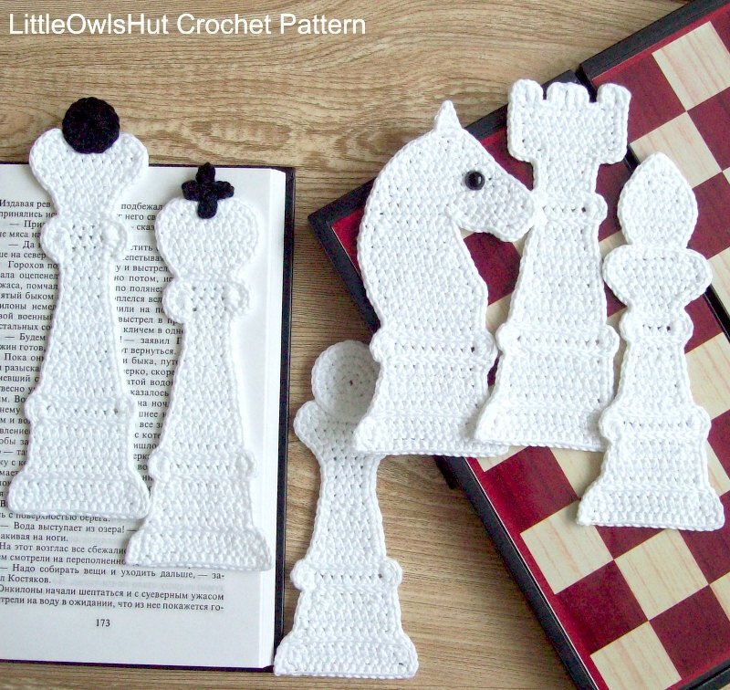 White crocheted chess pieces (king, queen, knight, rook, bishop, pawn) with some pieces marking pages in an open book and others placed next to a chessboard.