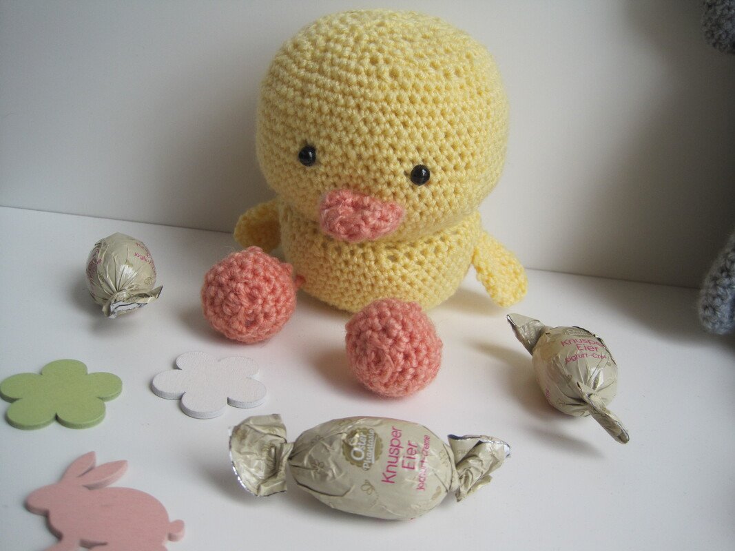 Gehäkeltes gelbes Amigurumi-Küken mit rosa Schnabel, losen rosa Häkelkugeln und eingewickelten Schokoeiern