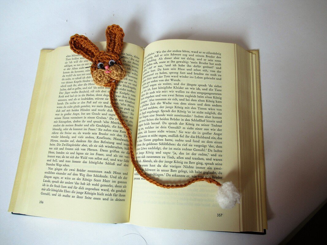 Gehäkeltes Hasen-Lesezeichen in Braun mit geflochtener Schnur und weißem Pompon auf offenem Buch