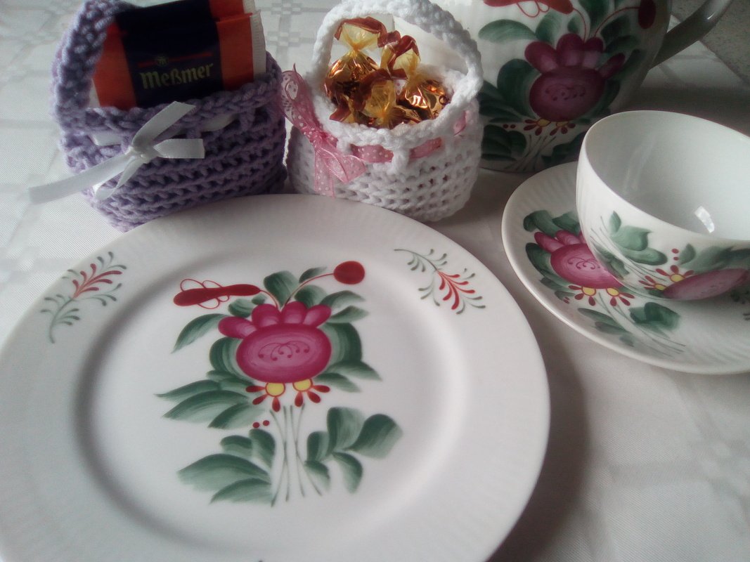 Zwei gehäkelte Mini-Körbchen (weiß und flieder) mit Schleifen, gefüllt mit Teebeuteln und Süßigkeiten, neben Teller und Tasse mit Blumenbemalung.