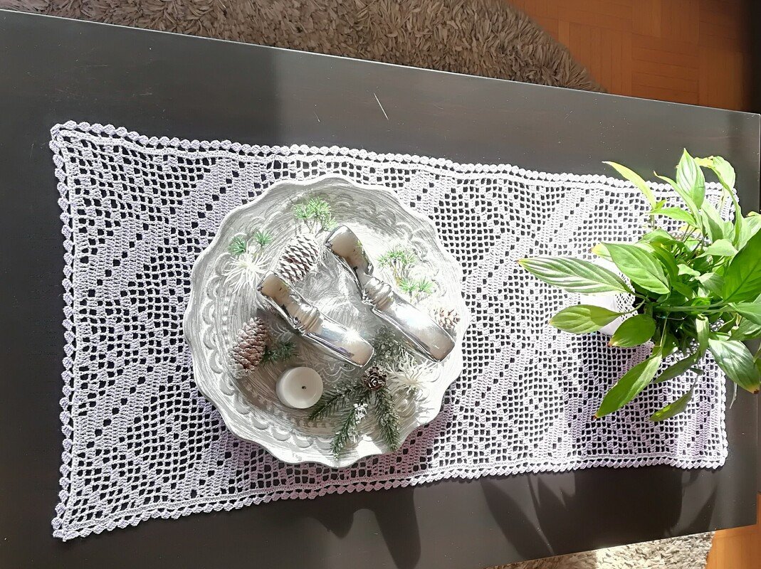 Weißer gehäkelter Tischläufer im Filetmuster mit Diamantmotiven auf dunklem Tisch, dekoriert mit Schale und Pflanze