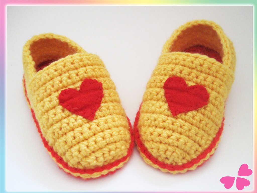 Gelbe gehäkelte Babyschuhe mit roten Herz-Applikationen und rotem Rand vor weißem Hintergrund.