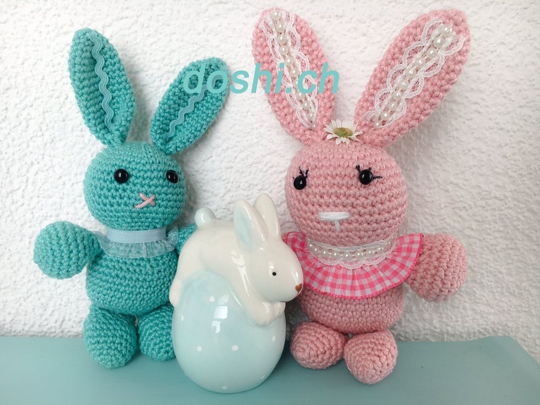 Zwei gehäkelte Amigurumi-Hasen, mintgrün und rosa, mit Perlendetails neben einer weißen Hasenfigur auf einem gepunkteten Ei.