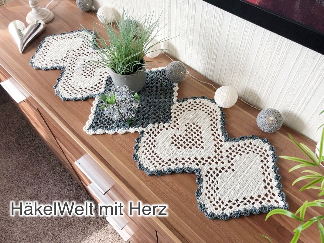 Gehäkelter Tischläufer aus Herz- und Quadratdeckchen in Weiß und Dunkelgrau auf einer Holzkommode, dekoriert mit Pflanzen und Lichterkette.