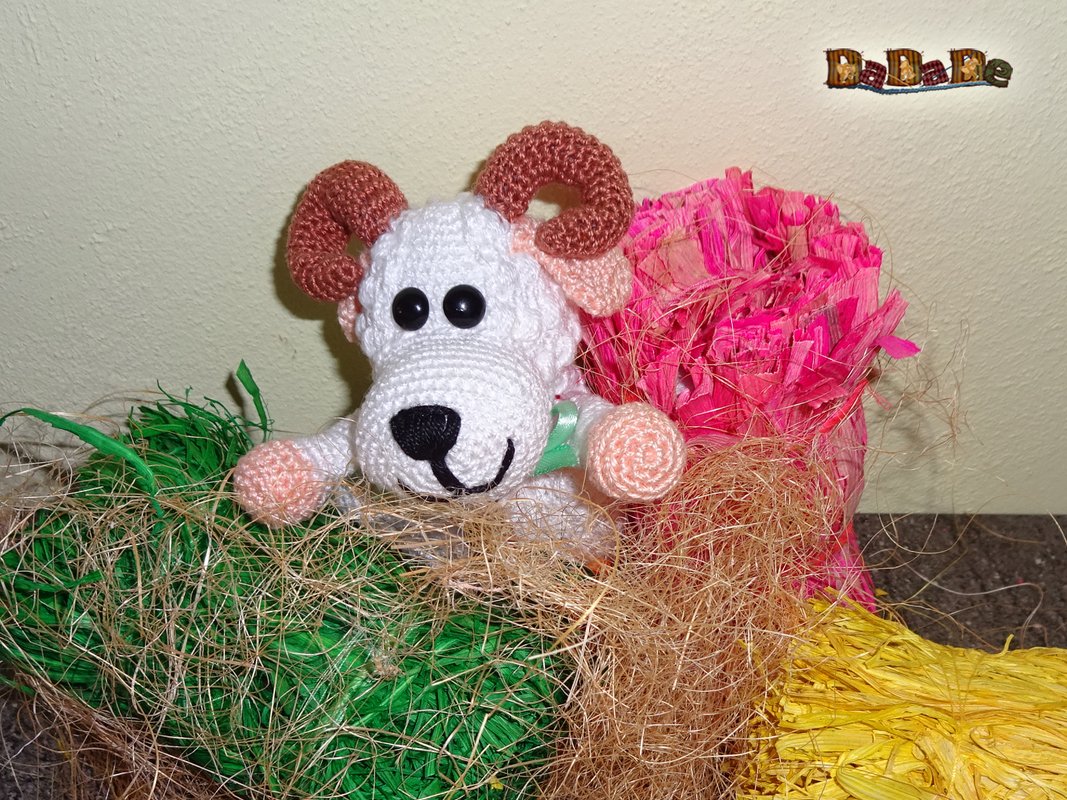 Weißes gehäkeltes Widder-Amigurumi mit braunen Hörnern auf mehrfarbigem Dekogras und Stroh