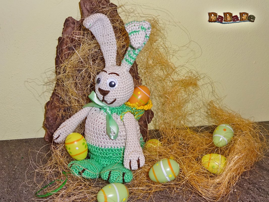 Gehäkelter Amigurumi-Osterhase in Beige und Grün mit kleinem Körbchen im Stroh, umgeben von bunten Ostereiern