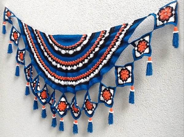 Blaues halbrundes gehäkeltes Tuch mit schwarzen, weißen und orangen Streifen; Rand aus Granny‑Squares mit blauen Quasten.