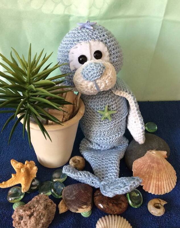 Gehäkelte hellblaue Amigurumi-Robbe mit Seestern-Applikation zwischen Muscheln und einer Zimmerpflanze