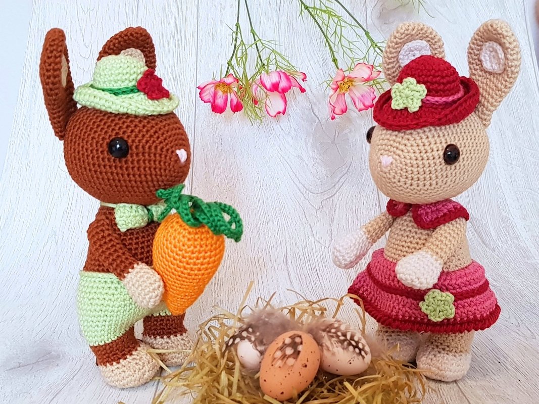 Zwei gehäkelte Amigurumi-Hasen mit Hüten, einer hält eine Möhre; davor ein Nest mit Ostereiern