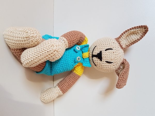 Schlafender Amigurumi-Hase in Beige und Braun mit türkiser Latzhose und gelbem Shirt auf hellem Hintergrund