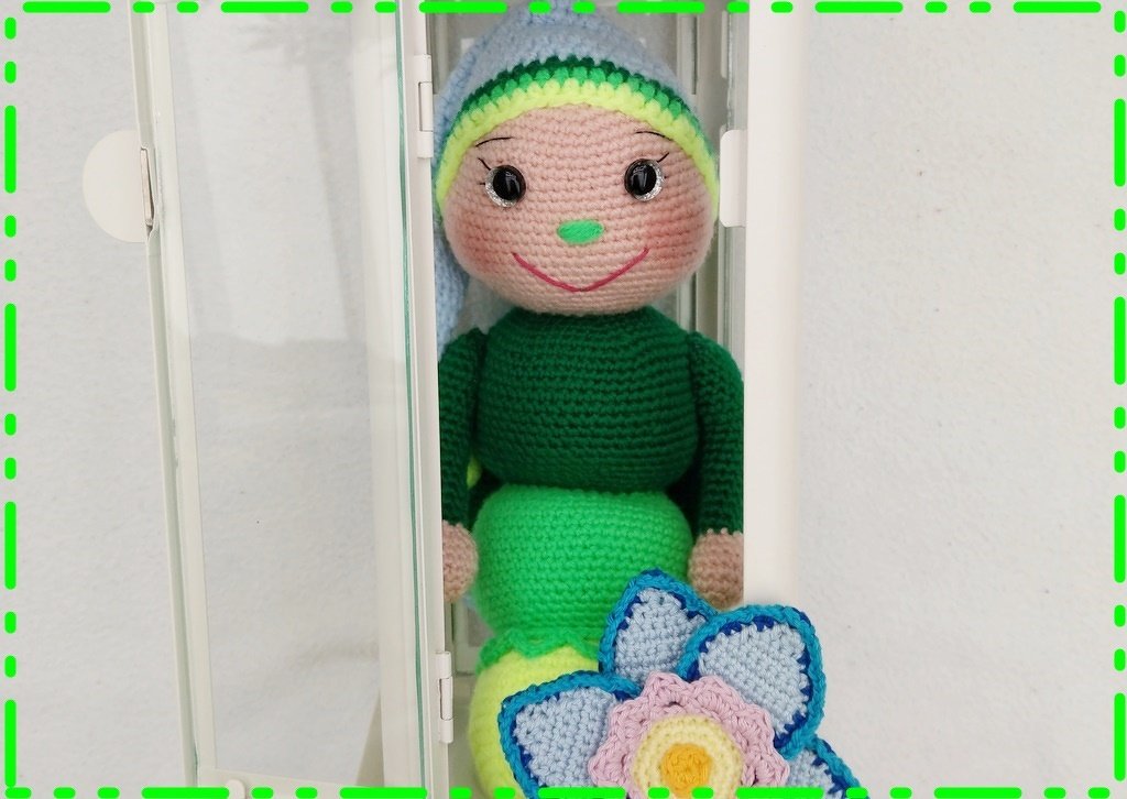 Gehäkelte Amigurumi-Puppe mit grünem Körper, blauer Zipfelmütze und gehäkelter Blume