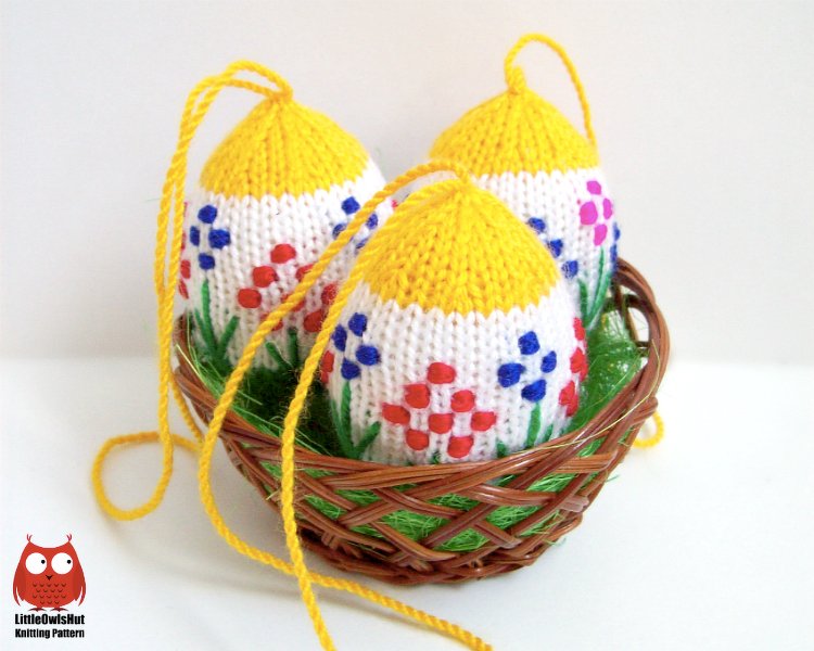 Three knitted Easter eggs with yellow tops and floral embroidery in a small wicker basket.