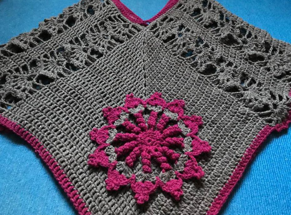 Grau gehäkelter Poncho mit pinker Blumenapplikation auf blauem Untergrund