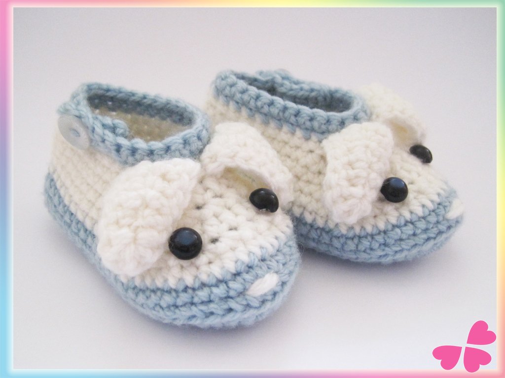 Hellblau-weiße gehäkelte Babyschuhe mit Hunde-Applikation und Knopfverschluss auf hellem Untergrund.