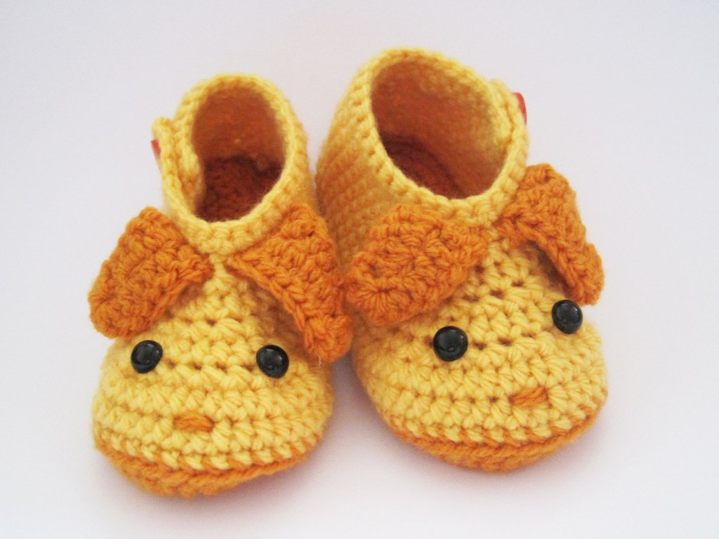 Gelbe gehäkelte Babyschuhe mit orangefarbenen Ohren und dunklen Knöpfen auf weißem Hintergrund