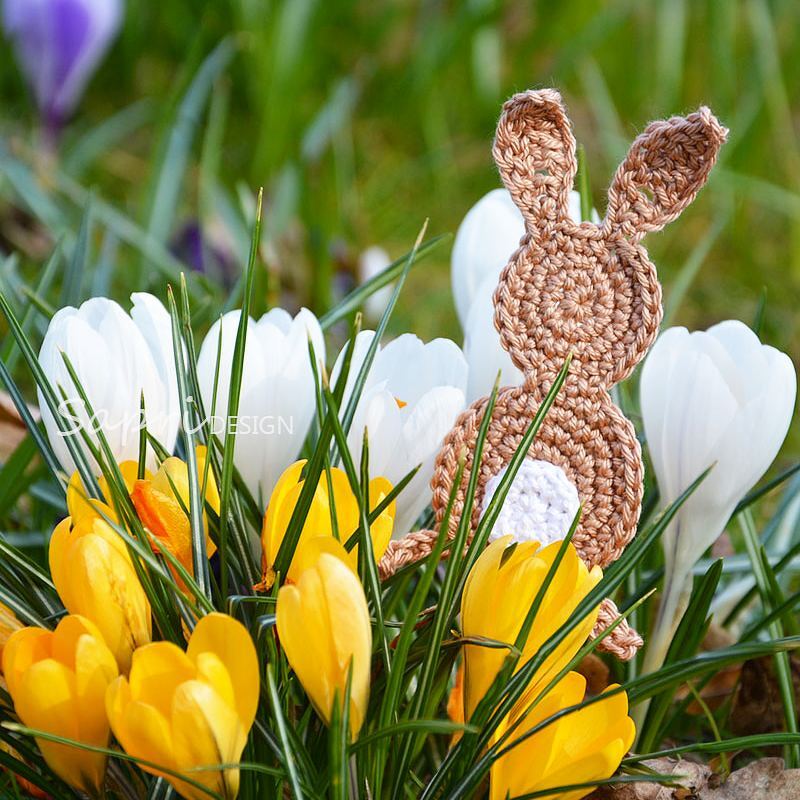 Gehäkelter Hase in Brauntönen mit weißem Schwanz, umgeben von gelben und weißen Krokussen im Gras.