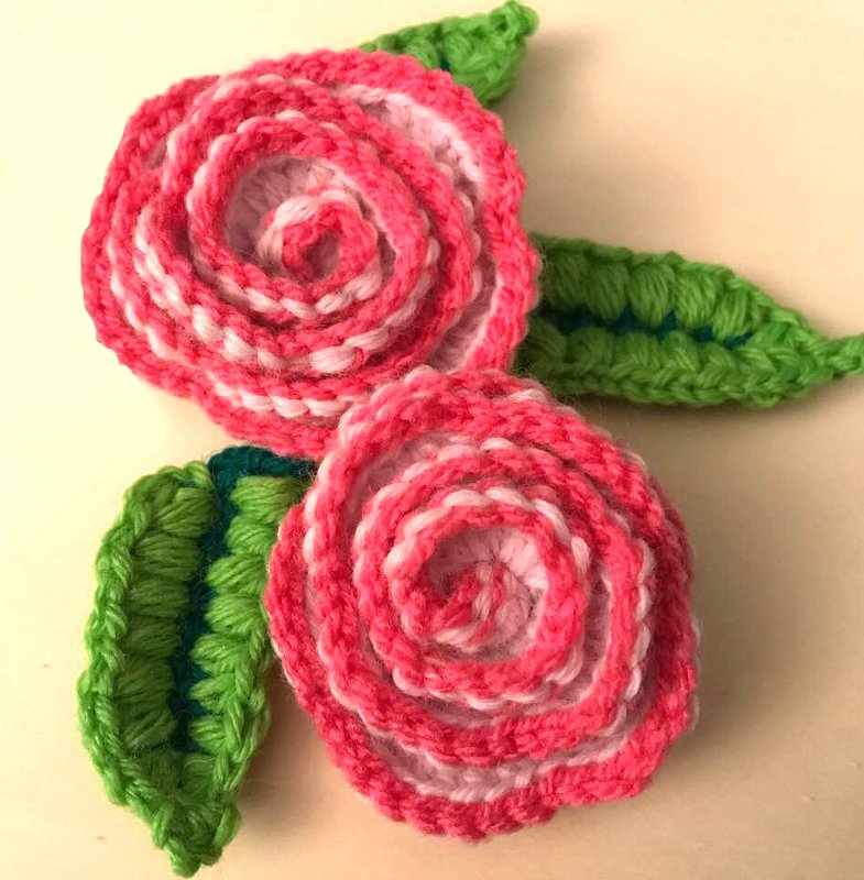 Zwei gehäkelte rosa-weiße Rosen-Applikationen mit grünen Blättern auf hellem Hintergrund.