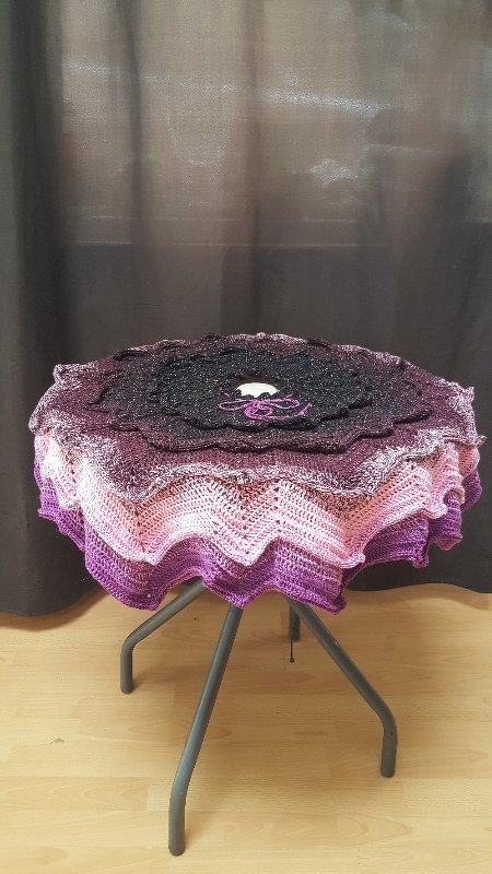 Runde gehäkelte Decke mit Farbverlauf in Schwarz, Lila und Pink auf dreibeinigem Hocker